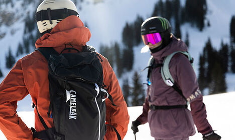 Two skiers at the top of a snowy mountain with hydration packs on.