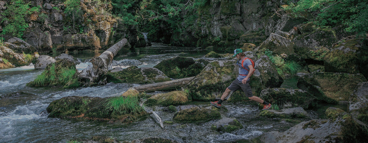 Man wearing hydration backpack stepping across stones in a river.