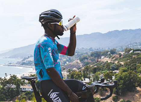 Cyclist resting on a mountain road drinking from a podium water bottle.
