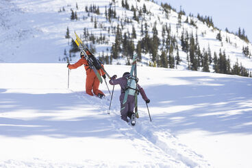 Two skiers walking up a snowy hill with their skis attached to snow packs.