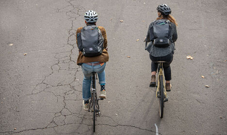Two bicyclists wear commute backpacks.