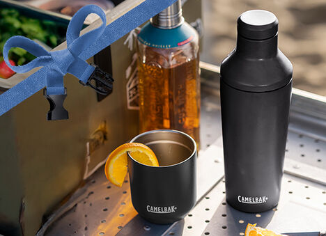 A Black Cocktail Shaker and rocks tumbler sitting on a camp bar with a bottle of whiskey and grill behind it.