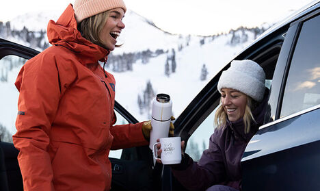 Person holding a customized camp mug while sitting in her car at the bottom of a snowy mountain.