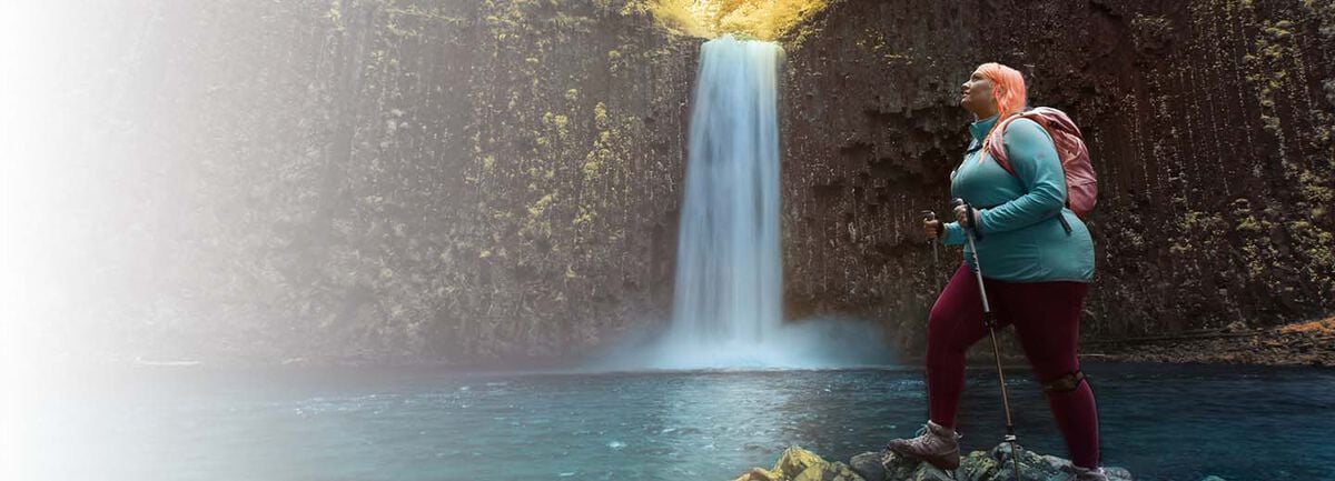 Jenny Bruso outside on a hike and standing on a rock with a waterfall in the background.