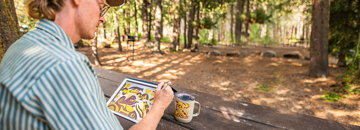 man wearing striped serch sitting at picnic table outside drawing design on ipad with mug nearby