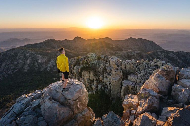 Mikah Meyer, in a yellow quarterzip, standing on top of a mountain in Big Bend National Park looking out at the view.