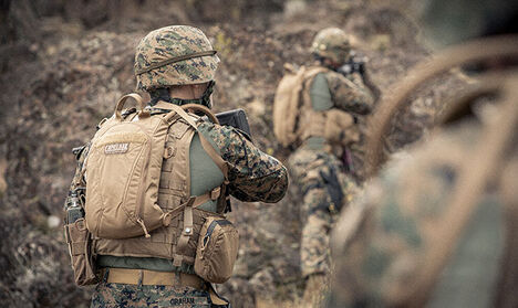 Two soldiers wearing hydration packs in a mountainous scene.
