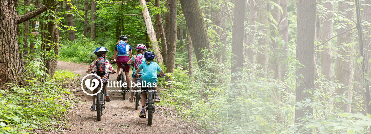 Women on bike leading 3 little girls on bikes on a trail in the woods.