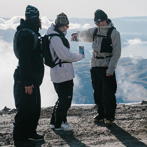 Three people sitting atop a mountain with drinks in hand and packs on their back.