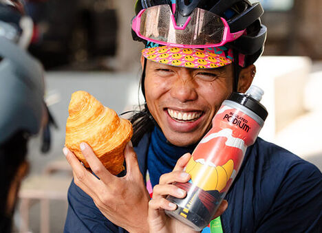 A cyclists holding a croissant in one hand and a Croissant Connoisseur Podium bottle in the other hand.