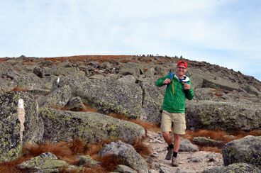 Mikah Meyer, in a green jacket and red hat, standing on a trail in Appalachian National Scenic Trail with a mountain view in the background.