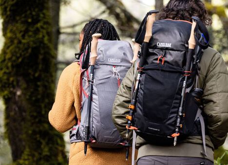 Two hikers going up a trail with smiles and hydration packs on.