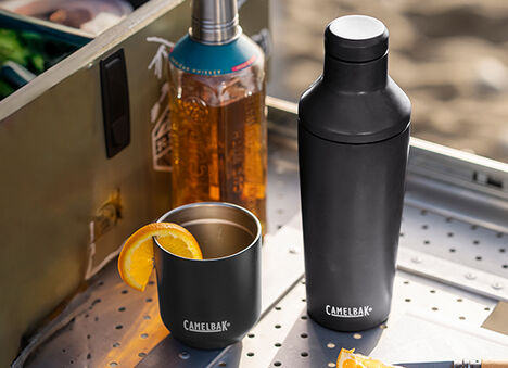 A Black Cocktail Shaker and rocks tumbler sitting on a camp bar with a bottle of whiskey and grill behind it.