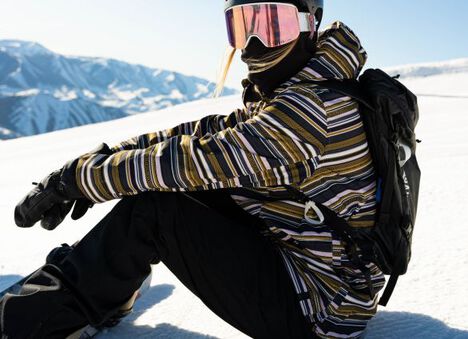 A snowboarder geared up at the top of the slope, glancing towards the camera before she goes down the mountain.