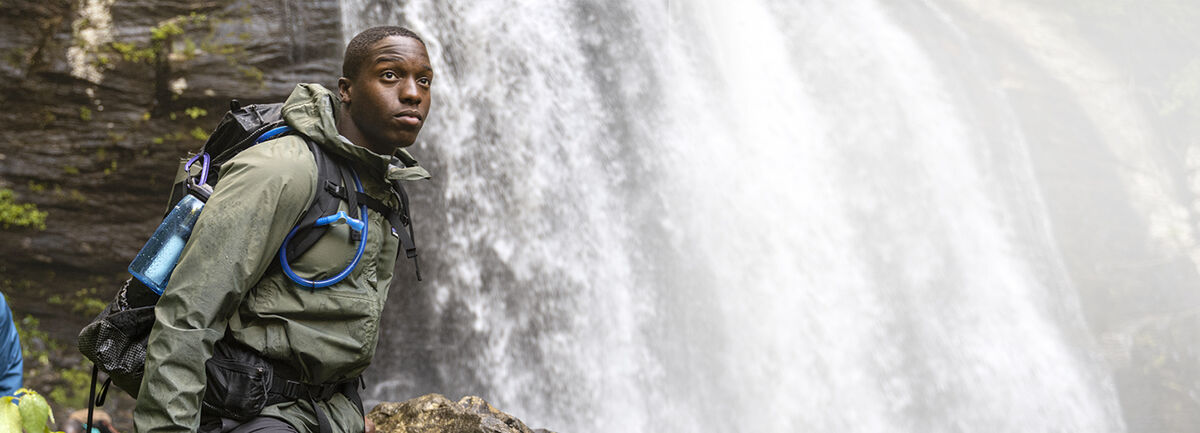 Man standing by waterfall carrying water bottle and hydration backpack.