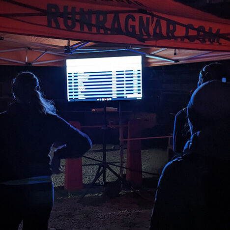 A few onlookers observing a screen which tells them when their team members are getting close to the handoff tent. 