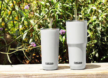 Two white Horizon stainless steel tumblers sitting on a railing with flowers behind them.