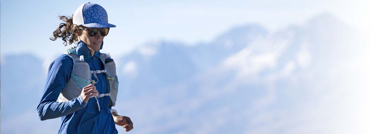 Women in a hat and wearing sunglasses running with mountains in the distance. 