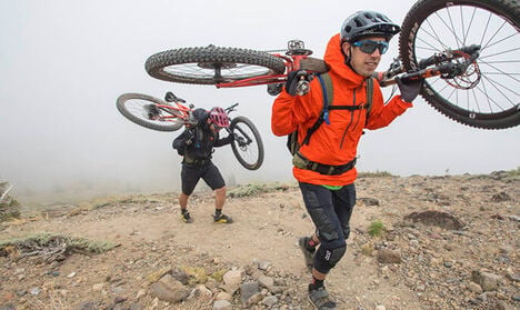 Two mountain bikers holding their bikes on their backs as they trek up a trail in the fog.