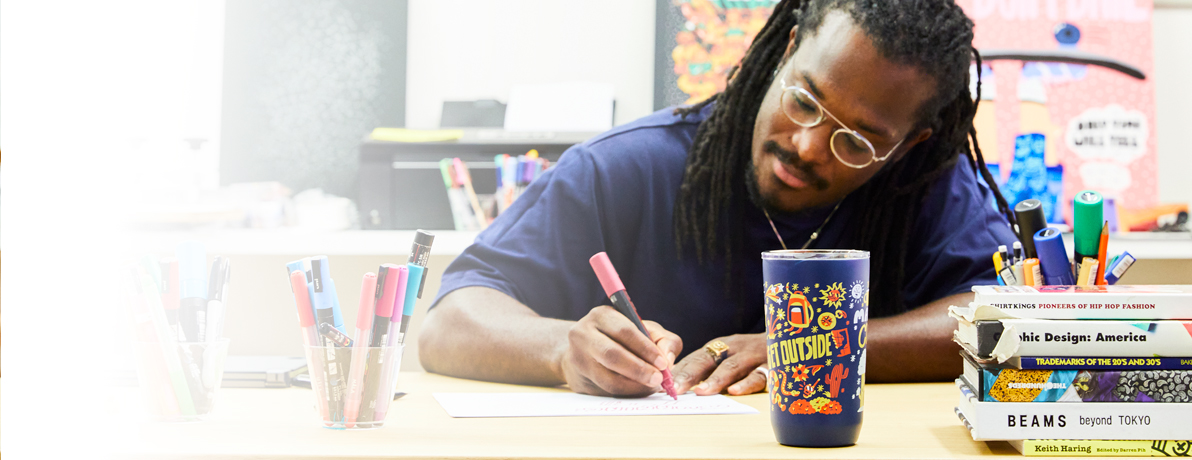 George F. Baker III sitting down at a desk drawing with a pink marker with his limited editon CamelBak vessel in front of him. 
