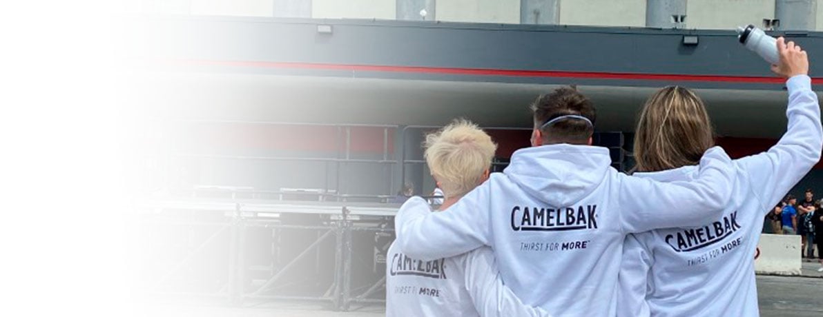 Three people facing away from the camera with arms around each other wearing white sweatshirts that have the CamelBak logo across the back. 