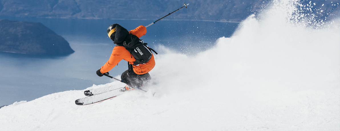 A person in a orange jacket skiing down a mountain wearing a CamelBak hydration backpack. 