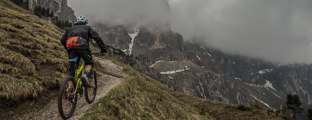 Mountain biker riding up steep trail toward view of cloudy mountains in distance.