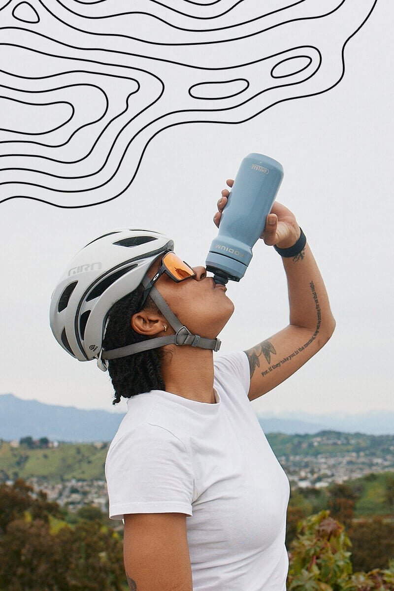 Biker drinking from a Podium Steel water bottle with a valley behind her and a topographical map overlay on the top of the image.