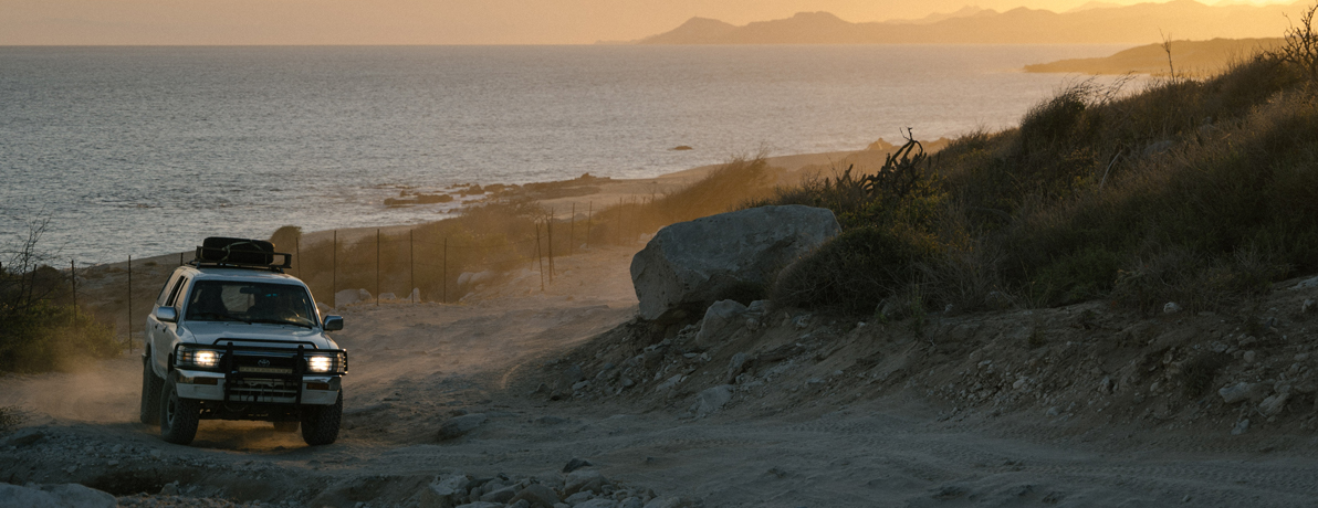 SUV driving on a dirt trail on the Baja coastline at dusk.