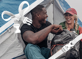 Two campers laughing as they sit in front of their tent, holding a fourteener hiking pack.