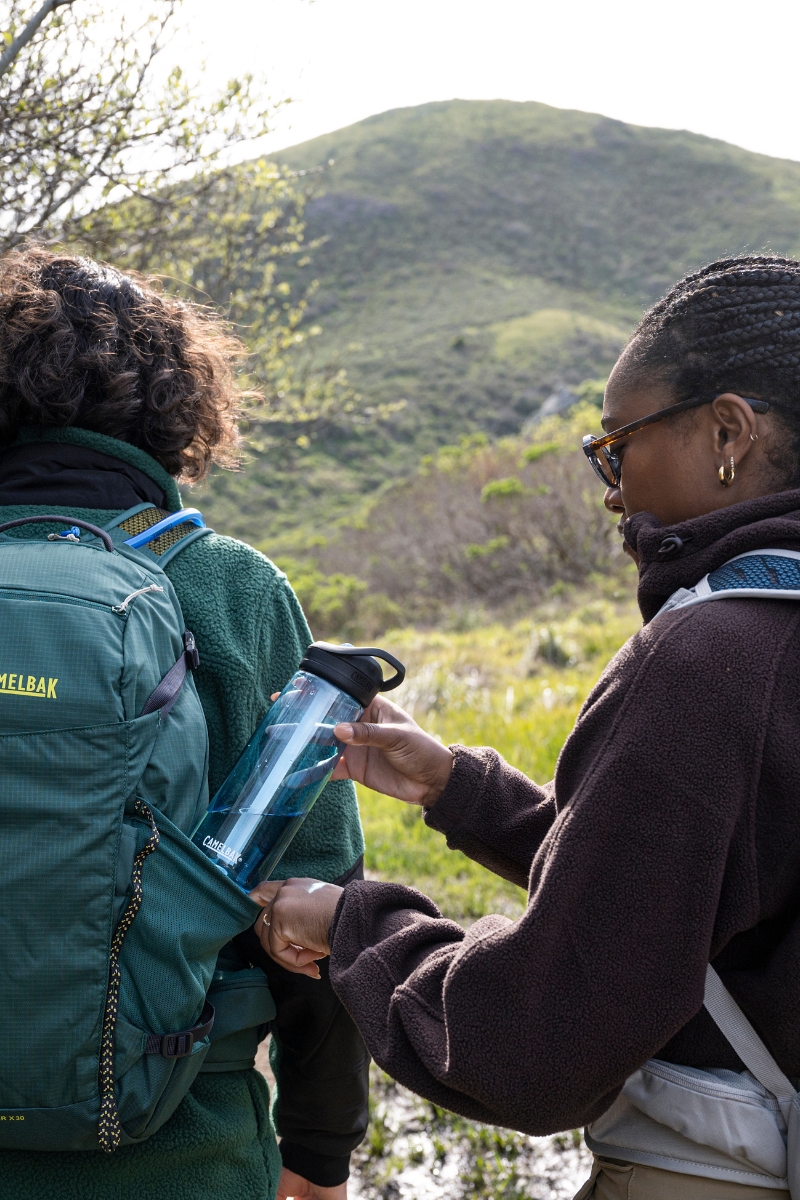 Two hikers stopping to grab a water bottle out of their hydration pack.