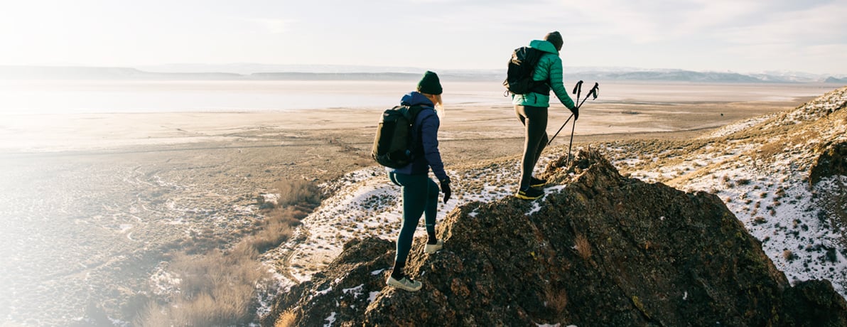 A man and a women in winter gear hiking up the side of a mountain that is dusted with snow.